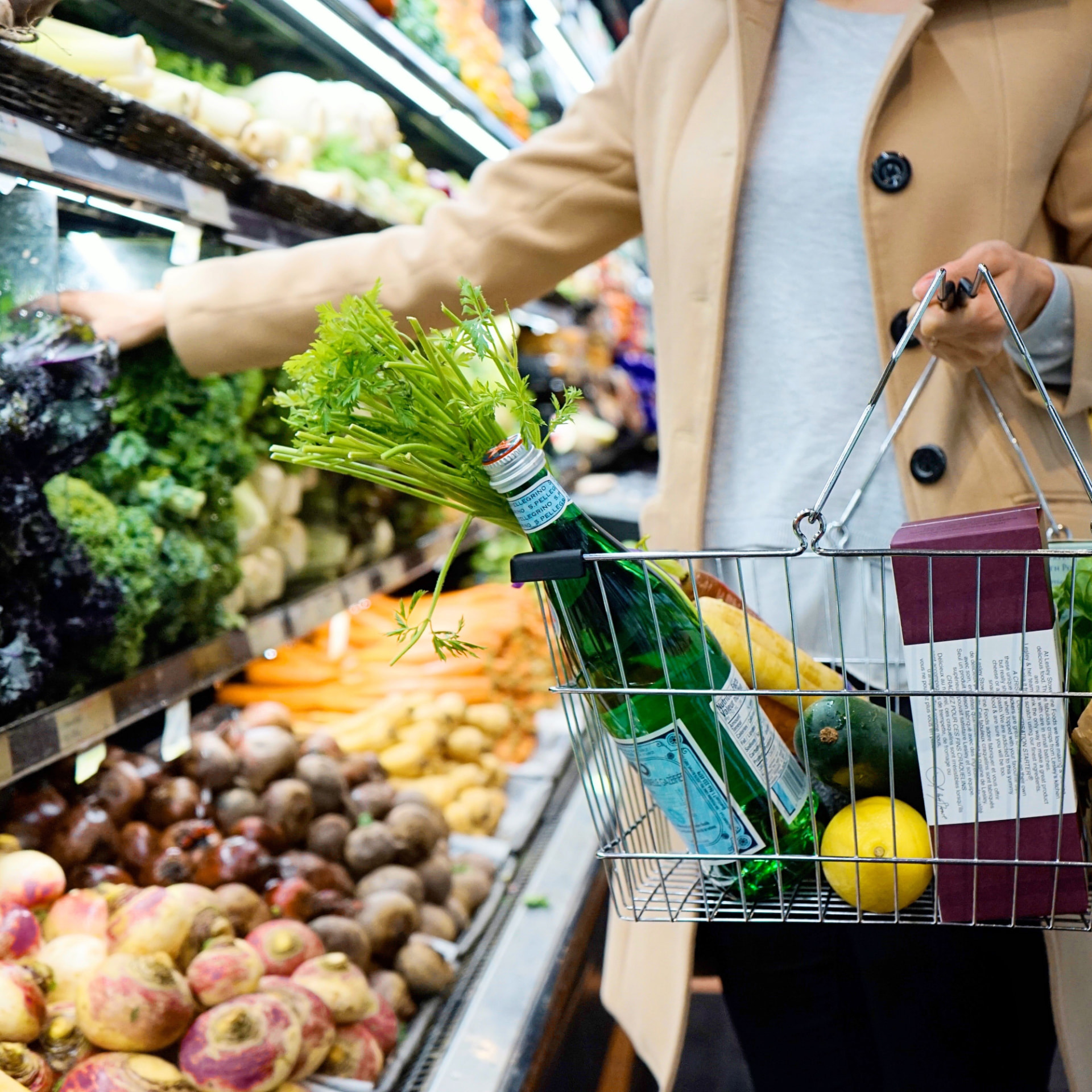 Shopper holding a basket with fresh vegetables, fruit and sparkling water in a supermarket produce aisle.